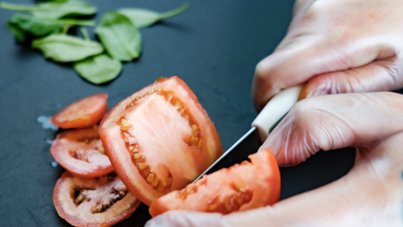 Chef's hands in food-safe gloves carefully slicing a fresh, ripe tomato on a dark cutting board, with basil leaves in the background, showcasing the use of fresh, local ingredients.