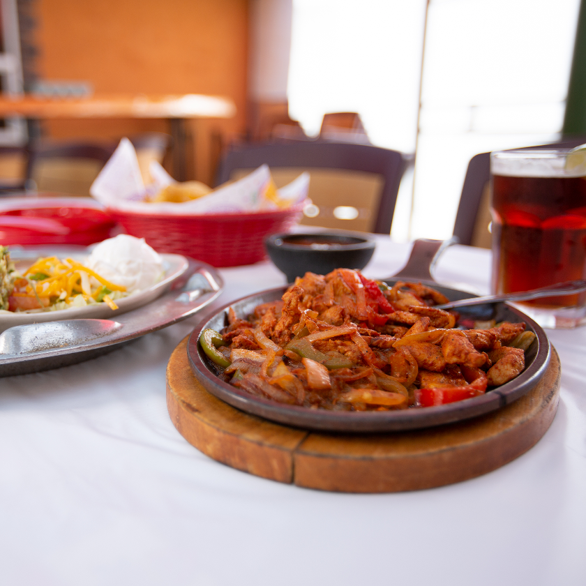 Sizzling fajitas on a wooden platter with accompanying side dishes, drinks, and table setting in a restaurant.